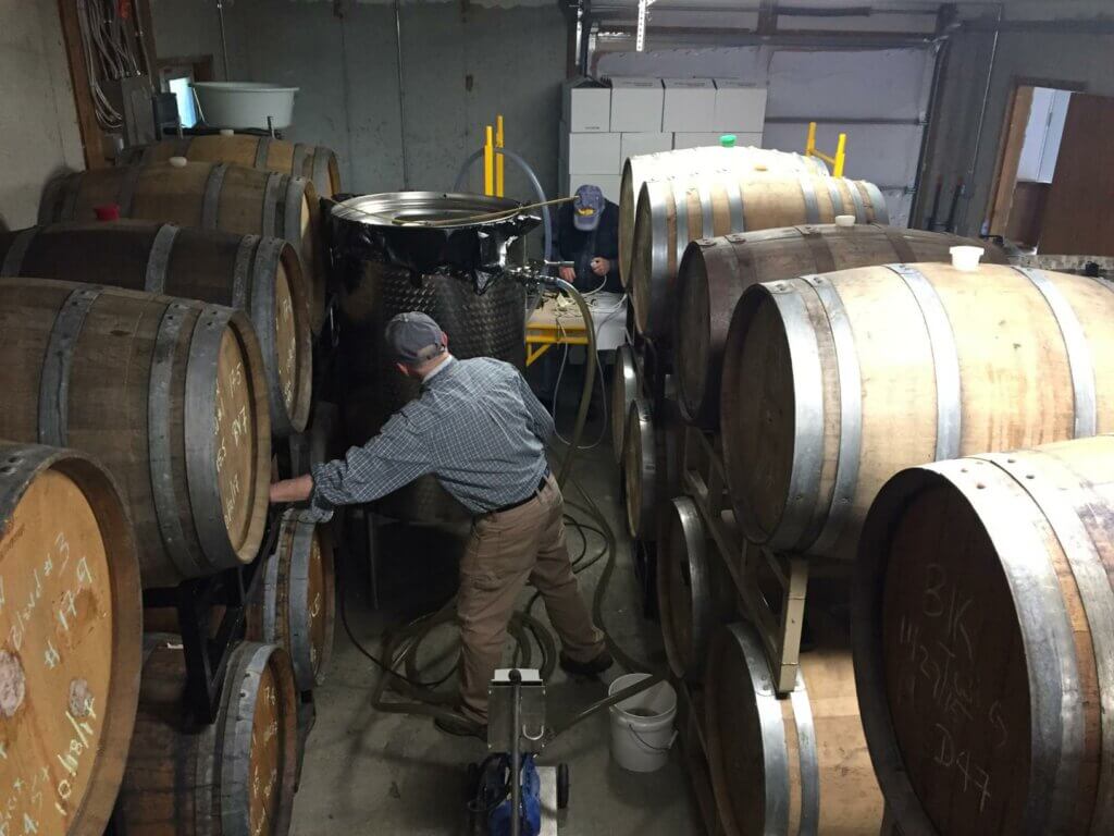 Two people in denim and ballcaps work in a room full of oak barrels, tubes and a large steel drum.