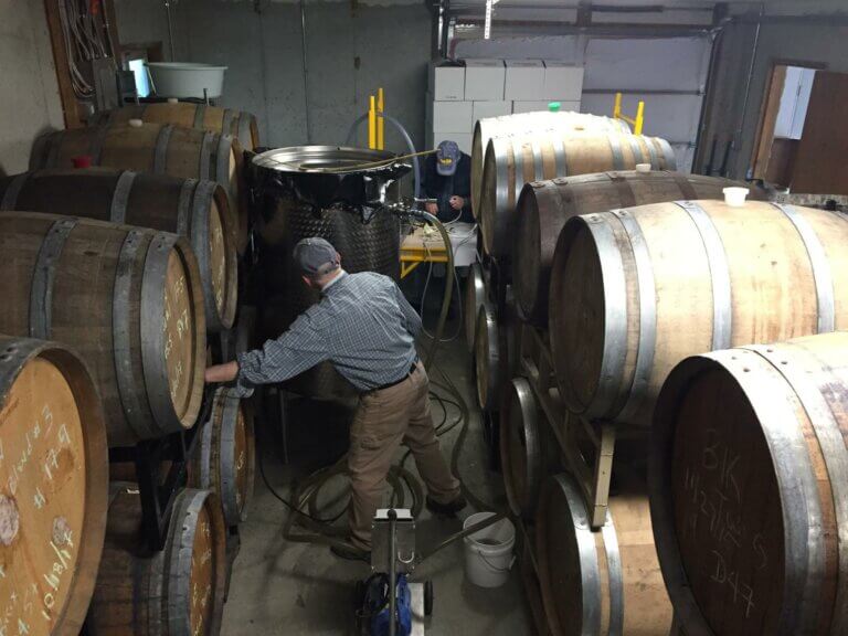Two people in denim and ballcaps work in a room full of oak barrels, tubes and a large steel drum.